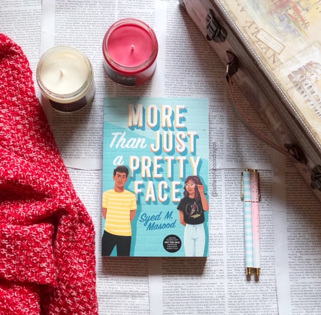 Picture of novel "More Than Just a Pretty Face' with two candles, a blanket, and suitcase in the sunlight