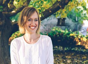 A woman with short blond hair and a white shirt is outside smiling at the camera