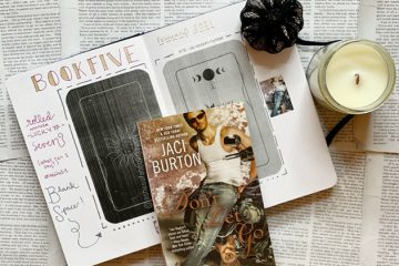 Paperback book of Don't Let Go by Jaci Burton reflecting a man in a white tank, leaning against a motorcycle with a yorkie dog at his feet. Paperback book rests on a journal with a candle off to the right side