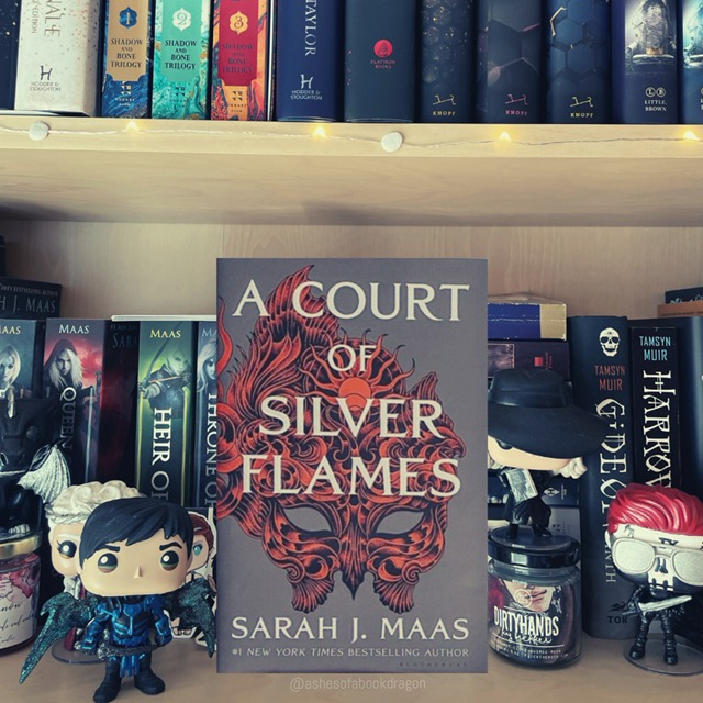 A Court of Silver Flames book resting on a bookshelf. The cover reflects an orange and black mask and the lettering for the title is white. 