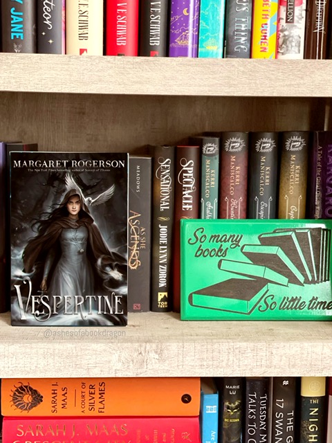 Bookcase stocked with books and a focus on a novel named "Vespertine" depicting a woman in a grey dress and dark colored robe. 