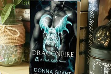 A paperback novel rests on a bookshelf. The book cover reflects a man's chest showing a light blue dragon tattoo. In front of the man and his tattoo, is the book name 'Dragonfire'.