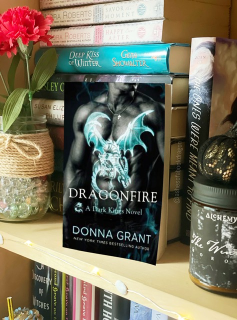 A paperback novel rests on a bookshelf. The book cover reflects a man's chest showing a light blue dragon tattoo
