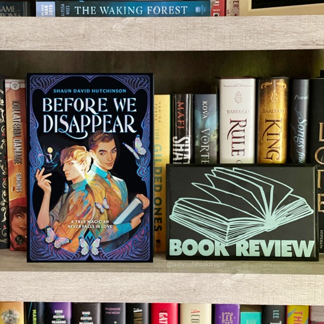 A book rests on a book shelf depicting two men on the cover, standing back to back. The man on the left has an arm raised with whisps of magic and the man on the right holds a book. 
