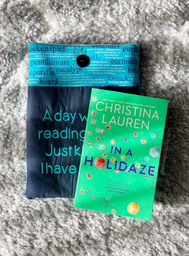 The book "In a Holidaze", light green with Christmas Tree ornaments on the over, rests on a book sleeve on a plush neutral background