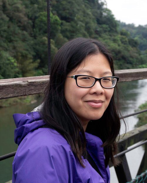 A woman with long dark hair, black glasses, and a purple coat smiles closed mouth at the camera 