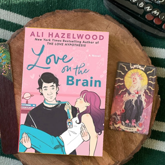 A pink book cover of "Love on the Brain" depicting a man with dark hair carrying a woman with pink and purple hair bridal style