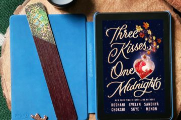 An e-reader rests on a wooden placemat with a bronze key, two candles, and a bookmark. The e-reader shows a book with the title "Three Kisses, One Midnight"