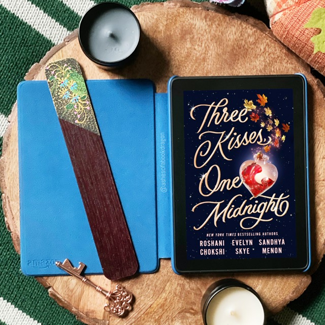 An e-reader rests on a wooden placemat with a bronze key, two candles, and a bookmark. The e-reader shows a book with the title "Three Kisses, One Midnight"